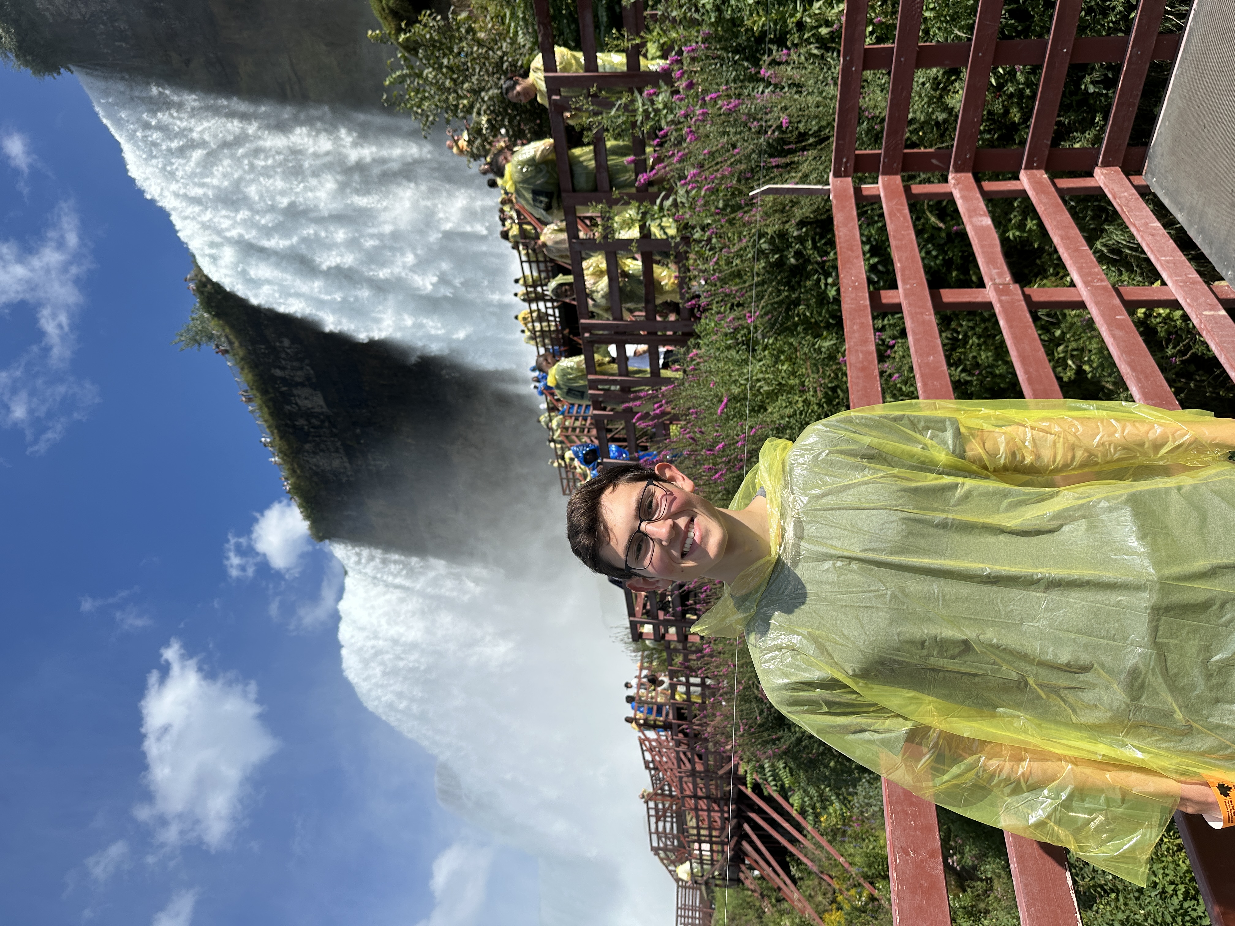 Justin in a bright yellow translucent rain pancho similing in front of niagra falls with purple flowers behind him on a board walk.